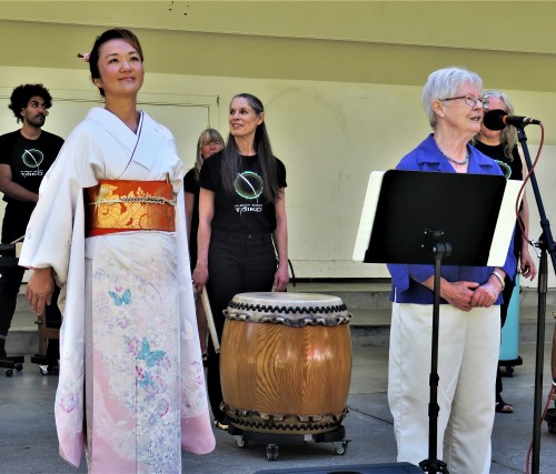 Miwa and Estelle stand on a stage in front of Taiko durmmers and address the crowd
